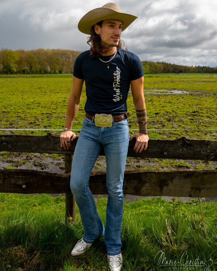Johnny Rossato in a cowboy hat leaning on a wooden fence in an open field